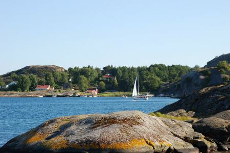 Klippor i förgrund med hav, en segelbåt och växtlighet i bakgrund.