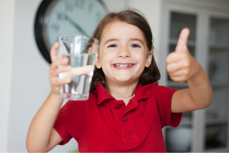 Barn i röd tröja håller upp ett glas vatten, ler stort och gör tummen upp.