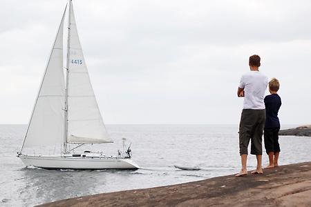 En vuxen och ett barn står på klipporna och en segelbåt seglar förbi.
