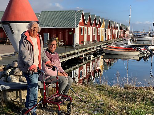 Två äldre personer framför småbåtshamn med sjöbodar.
